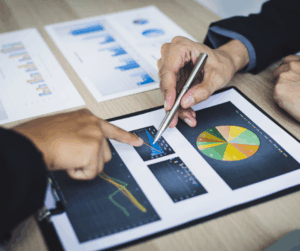 Hands pointing at charts and graphs on a clipboard and desk, representing data-driven decision-making for continuous improvement.