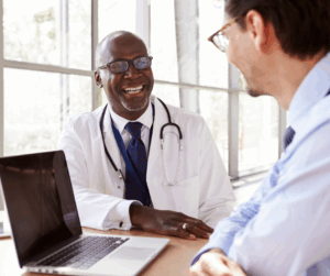Smiling doctor speaking with a patient across a desk with a laptop, representing secure communication and patient data protection.