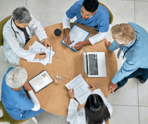 Overhead view of doctors and nurses in a meeting reviewing charts and data on paper, tablets, and a laptop, symbolizing coordinated care and secure patient information handling.