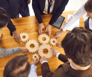 Group of people around a table connecting wooden gears, symbolizing teamwork and integrated secure systems.